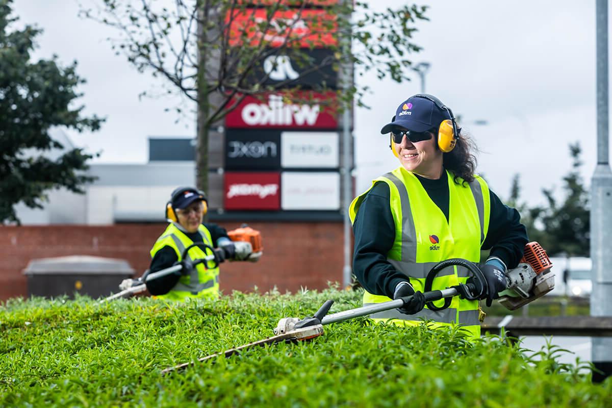 Two women wearing Mitie-branded high vis jackets, hats and safety glasses, strimming the top of a hedge